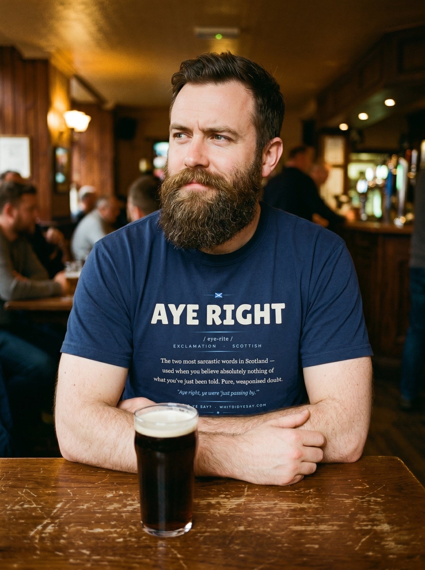 A man in a pub wearing the navy AYE RIGHT dictionary definition tee, with a pint on the table