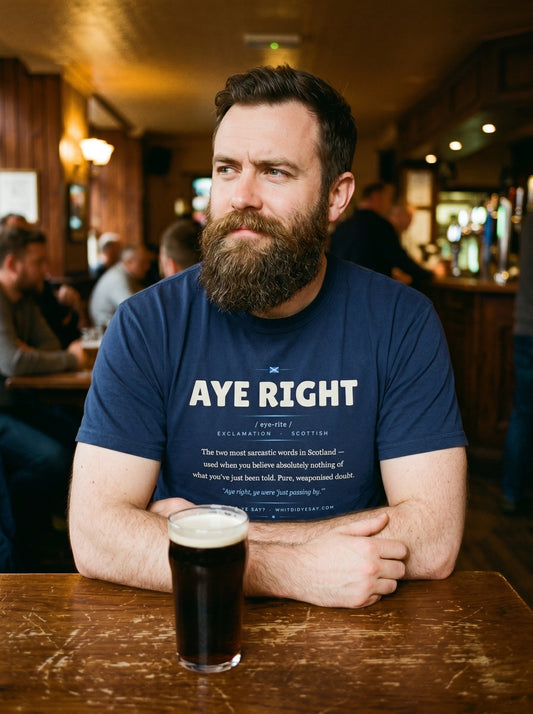 A man in a pub wearing the navy AYE RIGHT dictionary definition tee, with a pint on the table