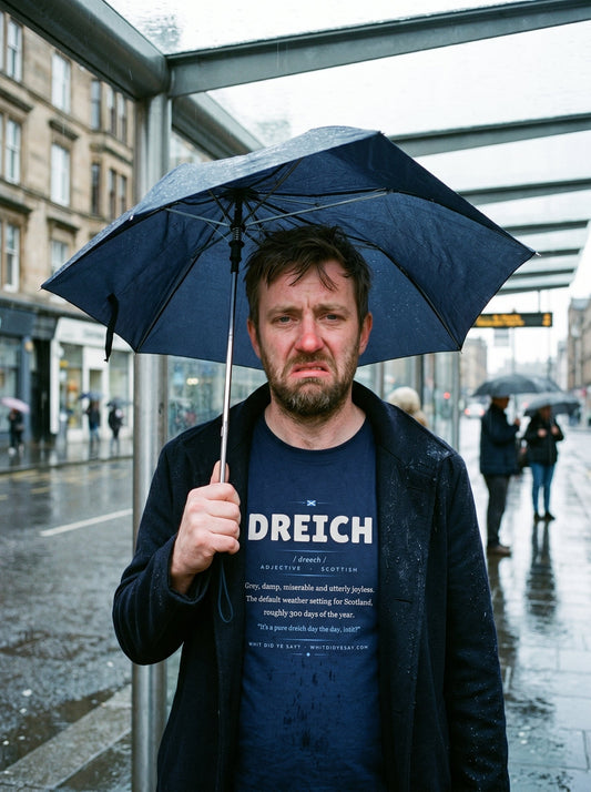 Man at a rainy bus stop with an umbrella wearing the navy DREICH dictionary definition tee